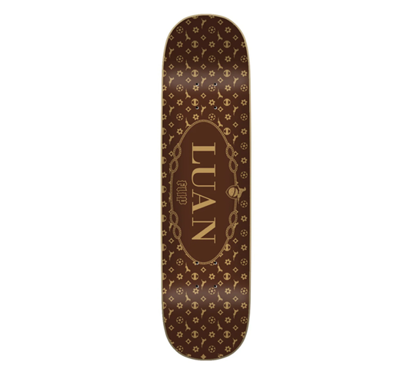 A skateboard deck shown from the top side, featuring a dark brown background with a repeating gold monogram pattern and a central gold oval logo that reads 'LUAN'.